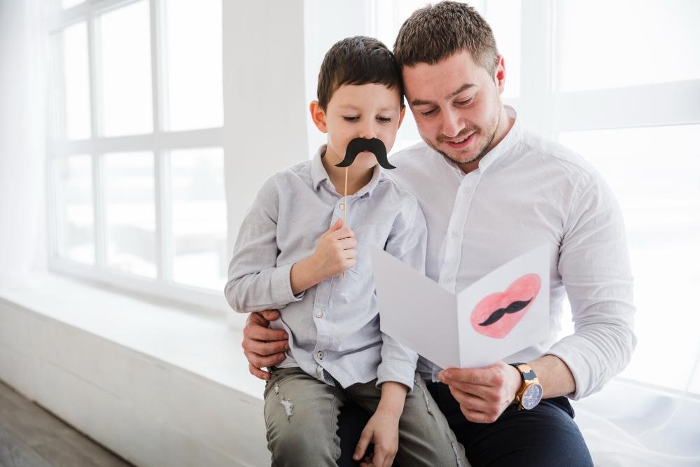 Um pai e seu filho se divertem juntos, com o pai lendo um cartão enquanto o menino segura um bigode de papel. Essa cena ilustra o tipo de pai que promove momentos de alegria e criatividade, fortalecendo o vínculo familiar através de brincadeiras e risadas.