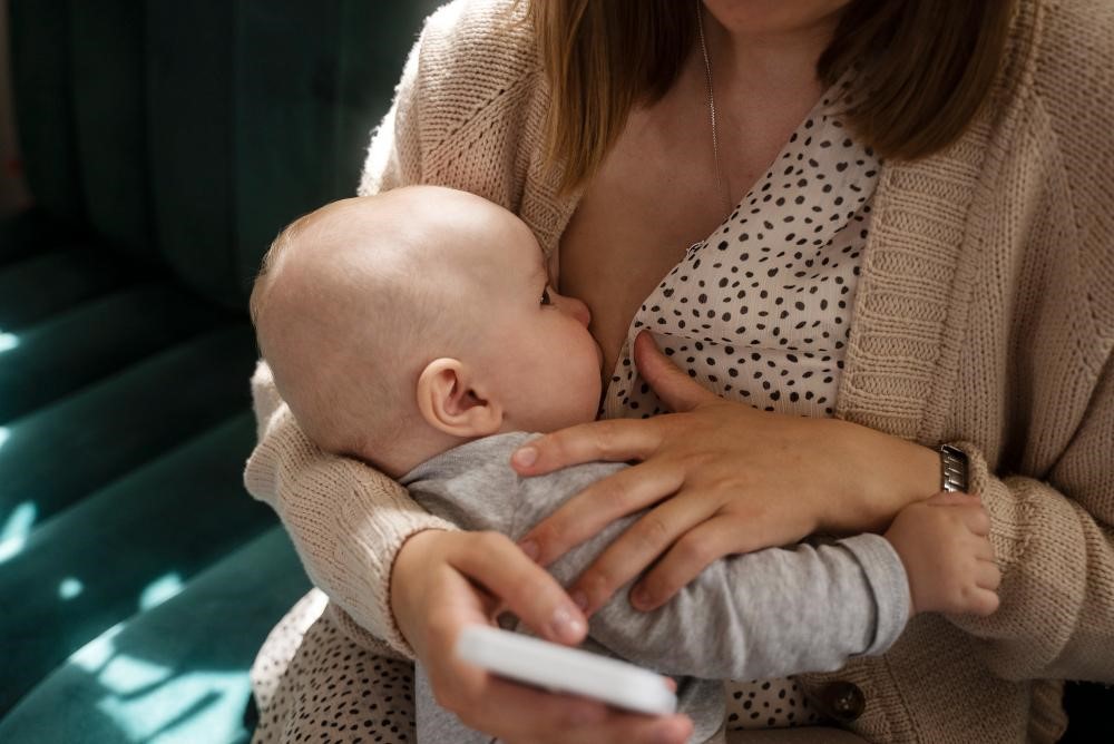 "Imagem de uma mãe praticando aleitamento materno, segurando seu bebê enquanto ele se alimenta. A mãe está vestindo uma blusa com estampa de bolinhas e um suéter, criando um ambiente acolhedor e íntimo para a amamentação.